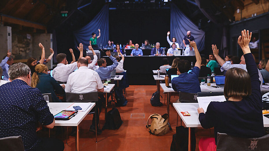 Bild von einer Parlamentssitzung, bei der gerade eine Abstimmung stattfindet und Parlamentarier:innen ihre Hände zur Abstimmung hochhalten.