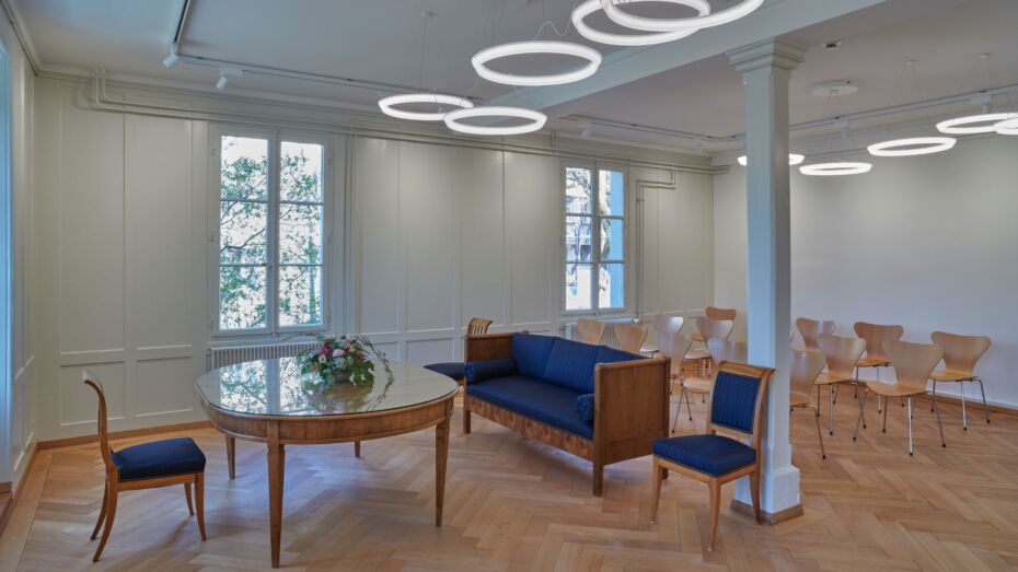 Ein ovaler Tisch, drei blau gepolsterte Stühle und ein kleines blau gepolstertes Sofa stehen dem Hochzeitspaar, den Trauzeugen und dem Standesbeamten zur Verfügung. Von der Decke hängen mehrere ringförmige Lampen und erzeigen ein angenehmes Licht. Im Hintergrund sind einfach Holzstühle für Hochzeitsgäste zu sehen.