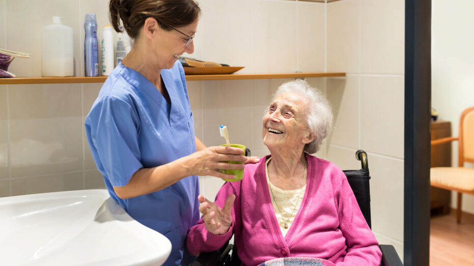 Eine jüngere, stehende Frau hilft einer älteren Frau im Rollstuhl beim Zähneputzen im Badezimmer.