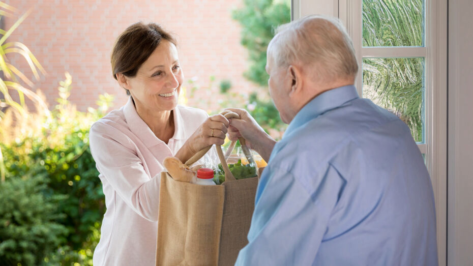 Eine Frau übergibt einem älteren, grauhaarigen Mann an der Haustüre eine mit Lebensmitteln gefüllte Einkaufstüte.